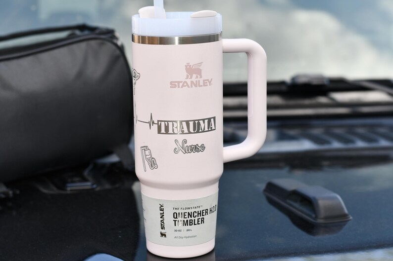 May include: A light pink Stanley Quencher H2.0 Flowstate tumbler with a white handle and lid. The tumbler has the word "TRAUMA Nurse" and a heartbeat graphic. The tumbler also has the Stanley logo and the words "Quencher H2.0 Tumbler" printed on it.