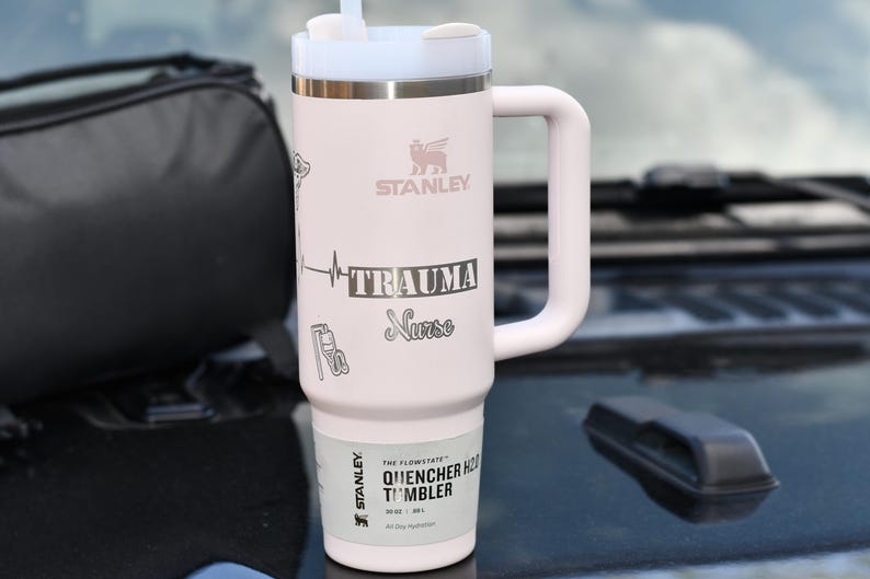 May include: A light pink Stanley Quencher H2.0 Flowstate tumbler with a white handle and lid. The tumbler has the words "TRAUMA Nurse" and a heartbeat graphic. The tumbler also has the Stanley logo and the words "Quencher H2.0 Tumbler" printed on it.