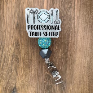 May include: A badge reel featuring a white acrylic topper with black line art of tweezers, scissors, a napkin, a scalpel, and shears. The text reads PROFESSIONAL TABLE-SETTER. Below are a turquoise glitter bead and a dark gray geometric bead.