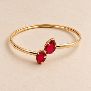 May include: A gold-toned open bangle bracelet featuring two pear-shaped, deep red faceted stones set in prong settings at the center. The metal band is smooth and circular, with the two stones positioned to face each other at the opening.