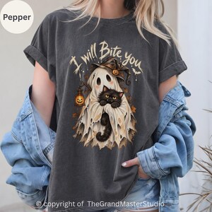 Könnte beinhalten: Ein anthrazitfarbenes T-Shirt mit einer Grafik eines Geistes, der einen Hexenhut trägt und eine Kürbislaterne hält, während eine schwarze Katze aus dem Geisterlaken hervorschaut. Der Text "I will Bite you" steht über der Geisterillustration.