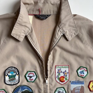 May include: Tan zip-up jacket with multiple embroidered patches. Patches include text and images of Colorado landmarks, such as Frisco, Emerald Lake, and Dream Lake. The jacket has a collared neckline.