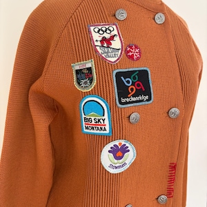 May include: An orange knit cardigan with multiple patches, including the Olympic rings and "Big Sky Montana." The cardigan has silver buttons and a ribbed texture. The patches feature various designs and text.