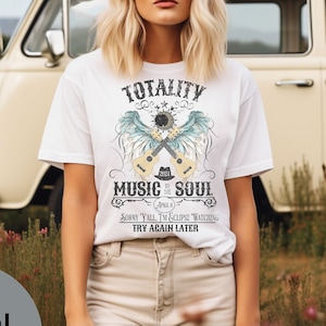 May include: White t-shirt with a graphic design featuring a pair of crossed guitars, wings, and the text "TOTALITY to the MUSIC SOUL" with the words "SORRY Y'ALL I'M ECLIPSE WATCHING, TRY AGAIN LATER" underneath.
