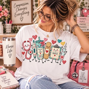 Könnte beinhalten: Ein weißes T-Shirt mit einer bunten Grafik aus medizinischen Cartoon-Utensilien, darunter ein Stethoskop, ein Thermometer, ein Becher mit der Aufschrift 'FUELED BY CARE', ein Pflaster, ein Klemmbrett mit 'NURSE' und eine Spritze, umgeben von Herzen.