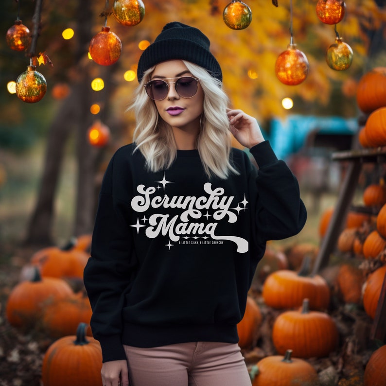 May include: Black sweatshirt with the text "Scrunchy Mama" in white script. The sweatshirt is paired with a black beanie and sunglasses. The background features pumpkins and decorative lights, creating a fall theme.