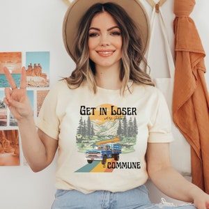 Może przedstawiać: Jasnożółta koszulka z vintage'owym nadrukiem niebieskiego samochodu ciężarowego jadącego górską drogą z tekstem "Get in Loser we're starting a commune".