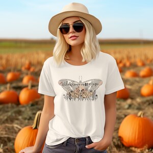May include: White t-shirt with a butterfly graphic and the text "Wildflowers by Design". The butterfly is outlined in black and has pink and orange accents. The shirt is worn by a person in a field of pumpkins.