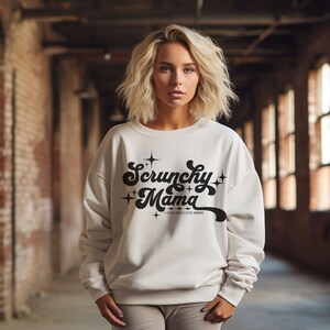 May include: Cream-colored sweatshirt with the text "Scrunchy Mama" in a retro font with star accents. The sweatshirt has a crew neck and long sleeves. The text "A little sexy & a little crunchy" is below the main text.