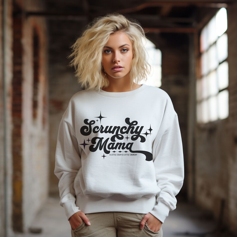 May include: A white sweatshirt with the text "Scrunchy Mama" in a black, retro font. The sweatshirt also has the text "A little silky & a little crunchy" below the main text. The sweatshirt is being worn.