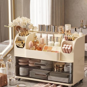 Pode incluir: Um organizador de maquiagem creme com vários compartimentos e gavetas. O organizador é preenchido com vários produtos de beleza, incluindo pincéis, batons, perfumes e produtos para a pele. Um arranjo decorativo de orquídeas fica no topo.