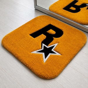 Puede incluir: Una alfombra cuadrada de color amarillo mostaza con esquinas redondeadas que presenta una letra R negra grande y una estrella negra con borde blanco. El diseño está centrado en la alfombra, que tiene una textura gruesa y afelpada.