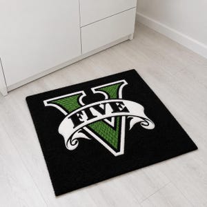 May include: A square black rug featuring a large green Roman numeral V with a textured scale pattern. A white banner is draped across the center of the V with the word FIVE printed in bold black capital letters, outlined in a thick white border.