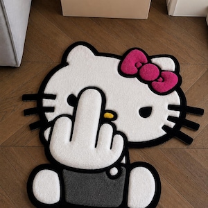 Puede incluir: Una alfombra de pelo cortado en blanco, negro y rosa con forma de personaje de gato de dibujos animados. El personaje tiene un lazo rosa, bigotes negros y una nariz amarilla. Una mano blanca muestra el dedo medio y viste una camisa gris.