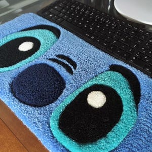 Puede incluir: Una alfombra rectangular con mechones que presenta un fondo azul claro con un rostro de personaje estilizado. El diseño incluye dos ojos grandes de color turquesa y negro con pupilas blancas, una nariz circular azul oscuro y dos cejas negras.