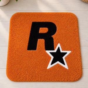 May include: A square tufted rug with a vibrant orange background featuring a large black letter R and a black star outlined in white. The rug has a thick, textured pile surface and rounded corners, displaying a bold graphic design on its front.