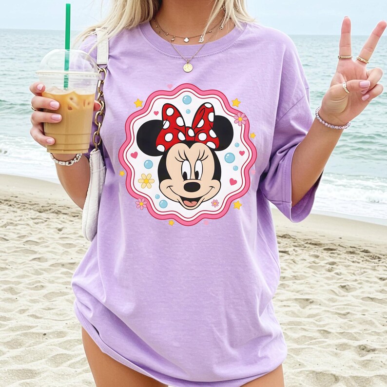 May include: A light purple t-shirt featuring a Minnie Mouse graphic. The graphic is a circular design with Minnie's face, a red bow with white polka dots, and a pink and white border. The shirt is worn on a beach.