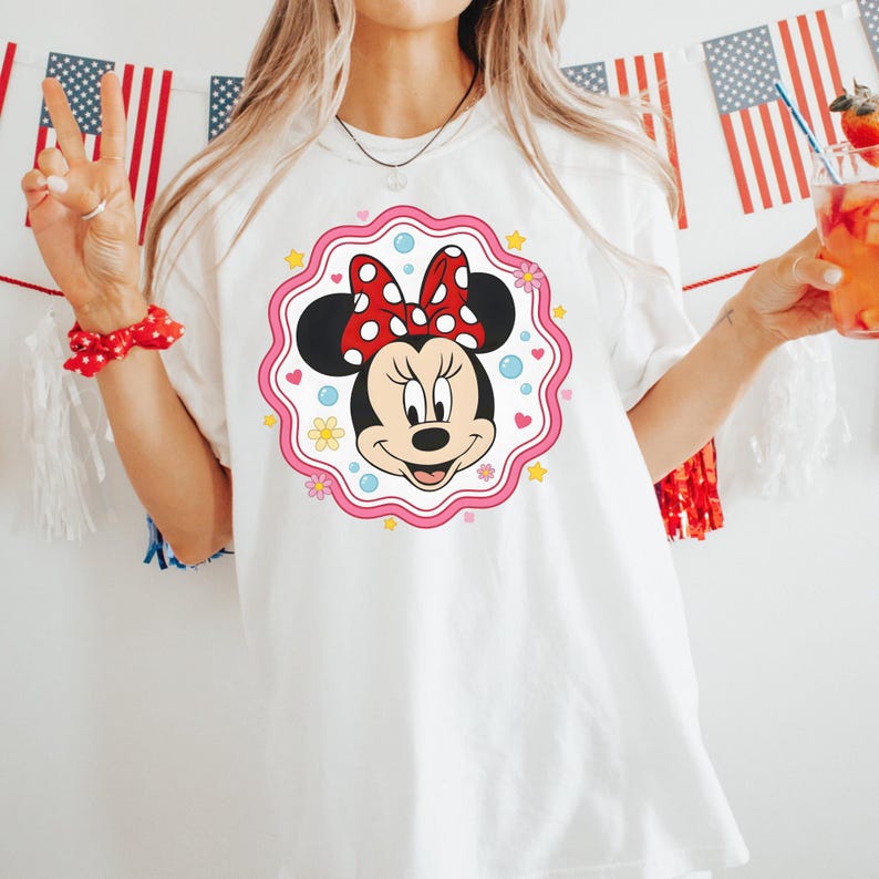 May include: White t-shirt featuring a Minnie Mouse graphic. The graphic includes Minnie's face, a red bow with white polka dots, and a pink scalloped border with flowers and stars. The shirt is a casual style.