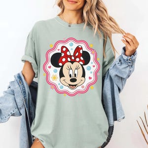 May include: A light green t-shirt featuring a Minnie Mouse graphic. The graphic shows Minnie's face with a red polka dot bow, surrounded by a pink and white scalloped border with flowers, hearts, and stars.
