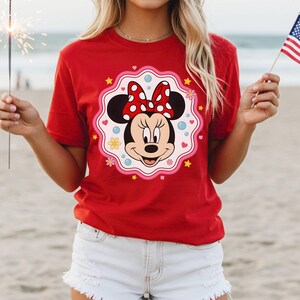 May include: A red t-shirt featuring a Minnie Mouse graphic. The graphic includes Minnie's face, a red bow with white polka dots, and a decorative border with hearts, flowers, and stars. The shirt is short-sleeved.