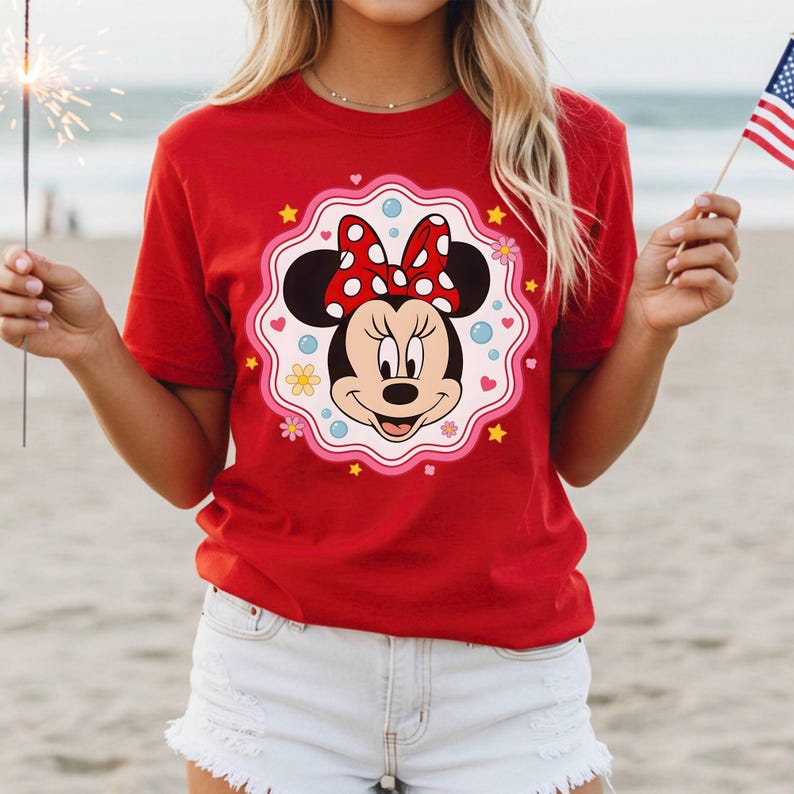 May include: A red t-shirt featuring a Minnie Mouse graphic. The graphic includes Minnie's face, a red bow with white polka dots, and a decorative border with hearts, flowers, and stars. The shirt is short-sleeved.