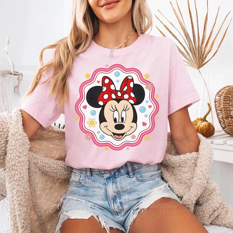 May include: A light pink t-shirt featuring a Minnie Mouse graphic. The graphic shows Minnie's face with a red bow with white polka dots, framed by a pink scalloped border with flowers and stars. The shirt is a casual wear item.