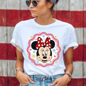 May include: White t-shirt featuring a Minnie Mouse graphic. The graphic includes Minnie's face, a red bow with white polka dots, and a pink scalloped border with stars and flowers. The shirt is worn by a person.