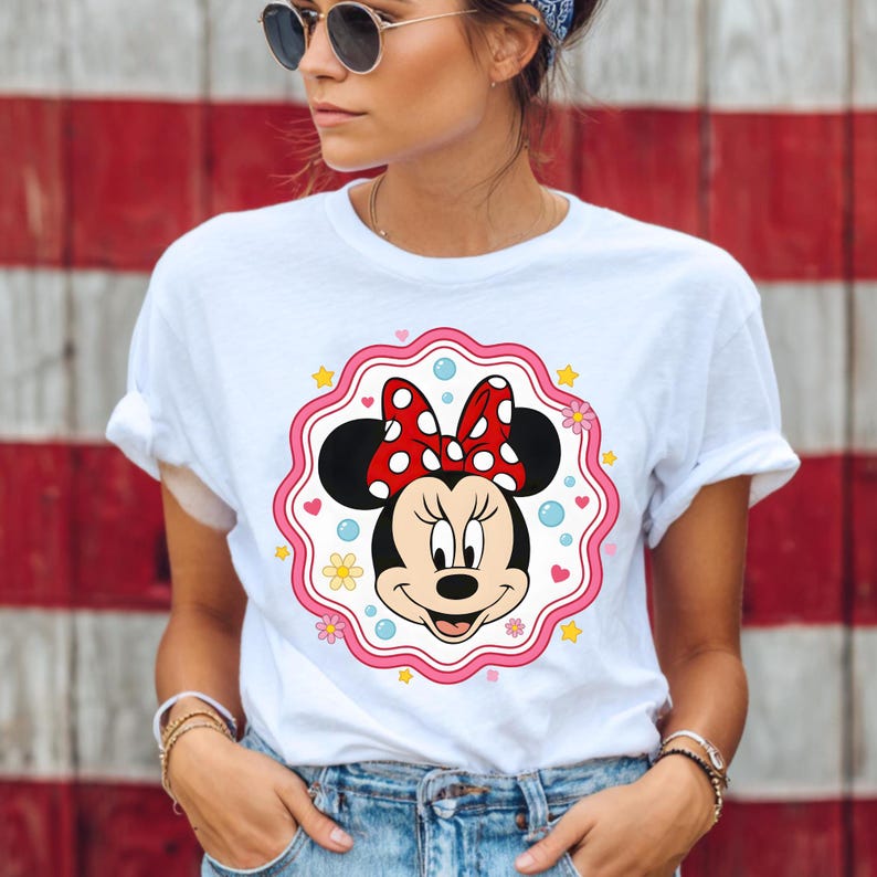 May include: White t-shirt featuring a Minnie Mouse graphic. The graphic includes Minnie's face, a red bow with white polka dots, and a pink scalloped border with stars and flowers. The shirt is worn by a person.