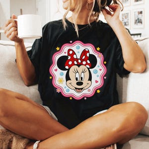 May include: Black t-shirt featuring a Minnie Mouse graphic. The graphic includes Minnie's face, a red bow with white polka dots, and a pink scalloped border with flowers, stars, and hearts. The shirt is a relaxed fit.