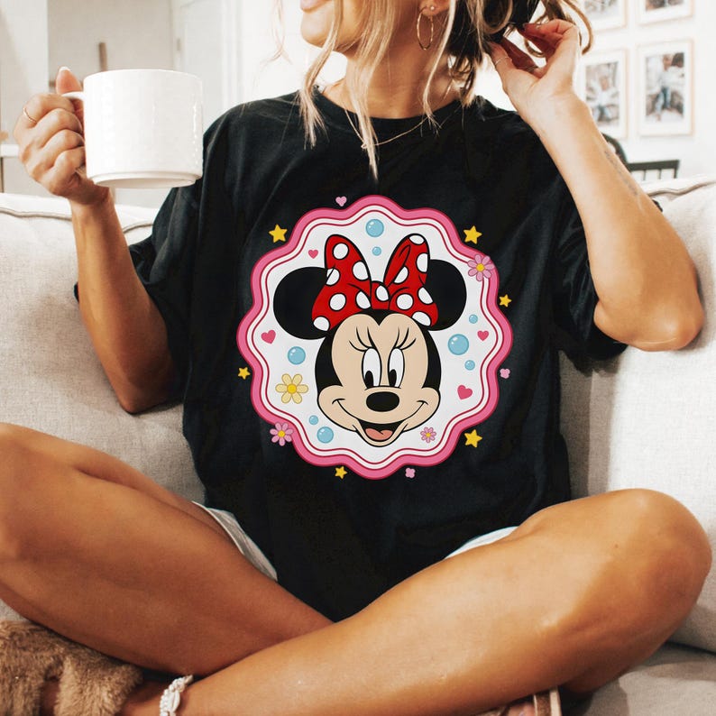 May include: Black t-shirt featuring a Minnie Mouse graphic. The graphic includes Minnie's face, a red bow with white polka dots, and a pink scalloped border with flowers, stars, and hearts. The shirt is a relaxed fit.
