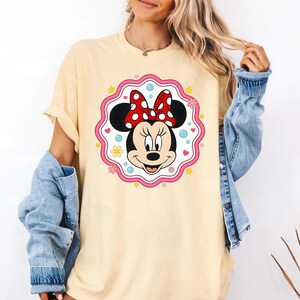 May include: A light yellow t-shirt featuring a Minnie Mouse graphic. The graphic includes Minnie's face, black ears, a red bow with white polka dots, and a pink and white scalloped border with hearts, stars, and flowers. The shirt has short sleeves.