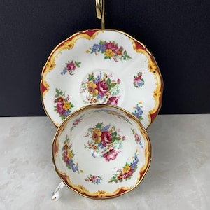 May include: A white porcelain teacup and matching saucer featuring a colourful floral pattern of pink roses, yellow flowers, and blue blossoms. The edges of both pieces are decorated with a scalloped gold trim and a deep red border design.