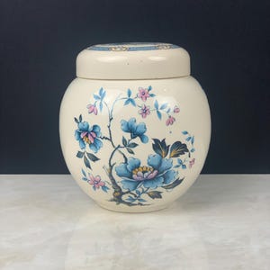 May include: A cream-coloured ceramic jar with a matching lid, featuring a decorative floral pattern of blue and pink blossoms with green leaves on the front. The jar has a rounded, bulbous shape and a smooth, glossy finish throughout the exterior.