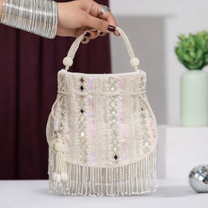 Puede incluir: Bolso de mano de cuentas marfil con asa y flecos de perlas. La bolsa presenta rayas verticales de lentejuelas y pequeños detalles de espejo. Una borla con detalles de perlas cuelga de un lado.