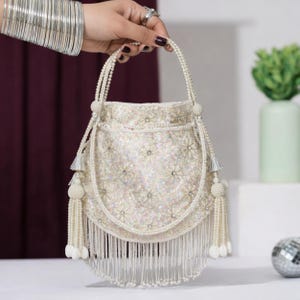 May include: A cream-colored drawstring handbag featuring an all-over sequined floral pattern with small embedded crystals. The bag includes a beaded pearl handle, decorative side tassels with pearl accents, and a long fringe trim along the bottom.