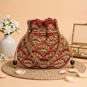 Könnte beinhalten: Ein tiefroter Samtbeutel mit Kordelzug, verziert mit aufwendigen goldenen und schwarzen Perlenstickereien in einem Bogenmuster. Die Tasche verfügt über geflochtene goldene Kordelgriffe mit dunkelbraunen Holzperlen an den Enden für ein strukturiertes Design.