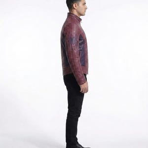 Peut inclure: Une veste en cuir bordeaux avec un motif &agrave; carreaux, un col montant et un embl&egrave;me d'araign&eacute;e bleu fonc&eacute; sur la manche. La veste est port&eacute;e avec un pantalon noir et des chaussures de ville en cuir noir, vue de profil.