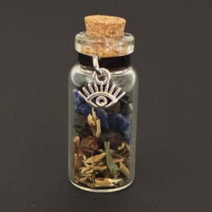 May include: A small clear glass vial with a cork stopper containing a mixture of blue stones, dark berries, and dried herbs. A silver-toned metal charm shaped like an eye is attached to the neck of the bottle with a black cord.