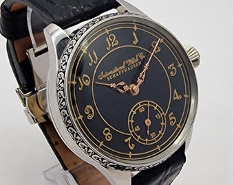 May include: A wristwatch with a black dial, gold-toned hands, and gold-toned Arabic numerals. The watch features a silver-toned case with an engraved pattern on the side and a black leather strap. The dial displays the text International Watch Co. SCHAFFHAUSEN.