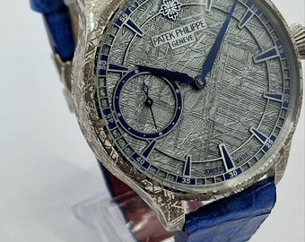 May include: A Patek Philippe Geneve wristwatch with a textured silver-toned dial featuring a cross-hatched pattern, blue hands, and a blue leather strap. The watch case has a rough, etched metallic finish and includes a small sub-dial.