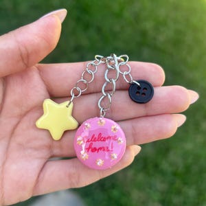 May include: A keychain featuring a silver metal chain with three charms: a pale yellow star, a round pink charm with the text Welcome home! in red script surrounded by small yellow dots, and a small black four-hole button attached to the chain.