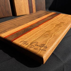 Op de afbeelding: Een rechthoekige houten snijplank met een lichtbruin oppervlak en een donkerbruine streep met een rode accent. De plank is gegraveerd met "The Smiths" en "Est. 2026".