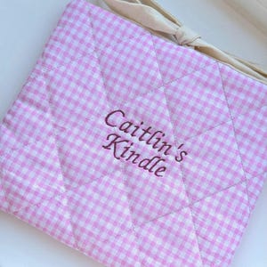 May include: A pink and white gingham Kindle cover with quilted stitching. It features a cream fabric tie and the embroidered text "Caitlin's Kindle" in a cursive font.
