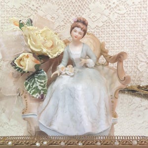 May include: A porcelain figurine of a woman in a blue and white dress sitting on a pink and white chaise lounge. The figurine is surrounded by a bouquet of yellow roses.