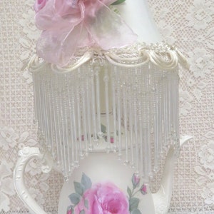 May include: A white ceramic teapot lamp with a pink rose design and a white lampshade with a pink ribbon and rose embellishments. The lampshade has a beaded fringe.