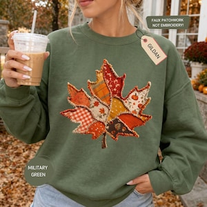 May include: A military green long-sleeve sweatshirt featuring a large central graphic of a maple leaf composed of various orange, red, and yellow autumn-themed fabric patterns. A Gildan tag is attached, and text on the image reads FAUX PATCHWORK NOT EMBROIDERY.