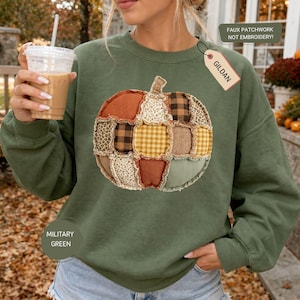 May include: A military green long-sleeve sweatshirt featuring a large central graphic of a pumpkin made of faux patchwork patterns in orange, brown, yellow, and cream. A GILDAN tag is attached, and text on the image reads FAUX PATCHWORK NOT EMBROIDERY.