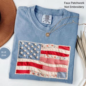 May include: A blue jean colored t-shirt with a faux patchwork American flag design. The flag features white stars, red and white stripes, and floral patterned fabric accents. A gold necklace with a pendant rests on the shirt.