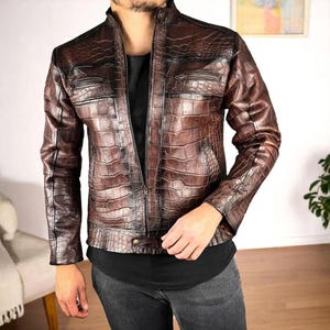 Peut inclure: Veste en cuir marron avec une texture peau de crocodile. La veste a une fermeture éclair, deux poches poitrine et deux poches latérales. La veste est de couleur marron moyen.