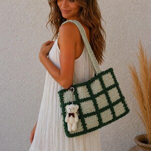 May include: A green and cream crochet tote bag with a square pattern and a matching strap. A small white teddy bear charm with a red bow hangs from the bag. The bag is held by a person wearing a white dress.